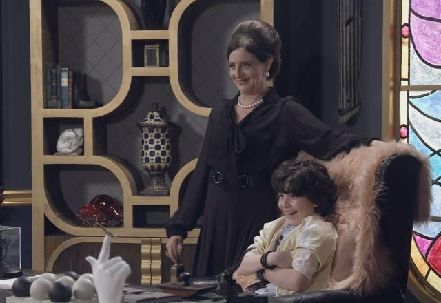 Norma, em pé, sorri ao lado de Felipe, que está sentado na cadeira da diretora, também sorrindo
