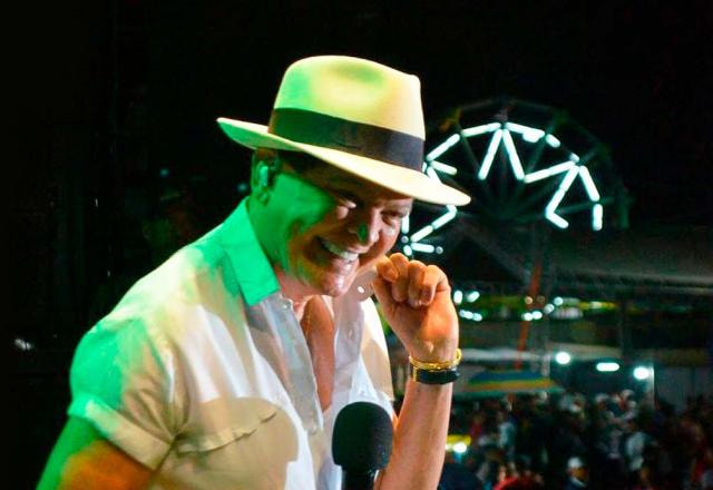 Alcymar Monteiro está com o microfone na mão. Ele veste branco e tem um chapéu Panamá na cabeça