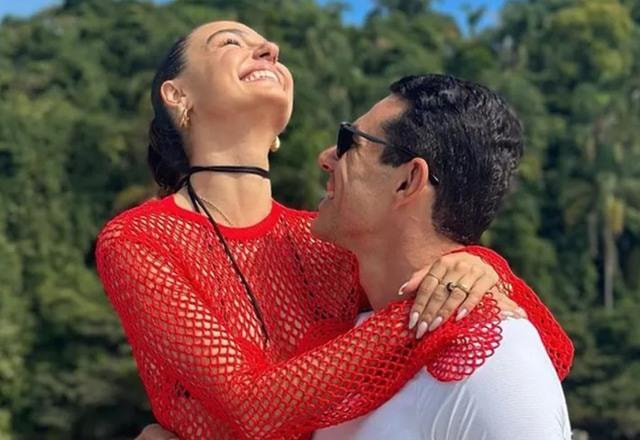 Foto de Isis Valverde sendo abraçada por seu noivo Marcus Buaiz, estida de vermelho e sorrindo